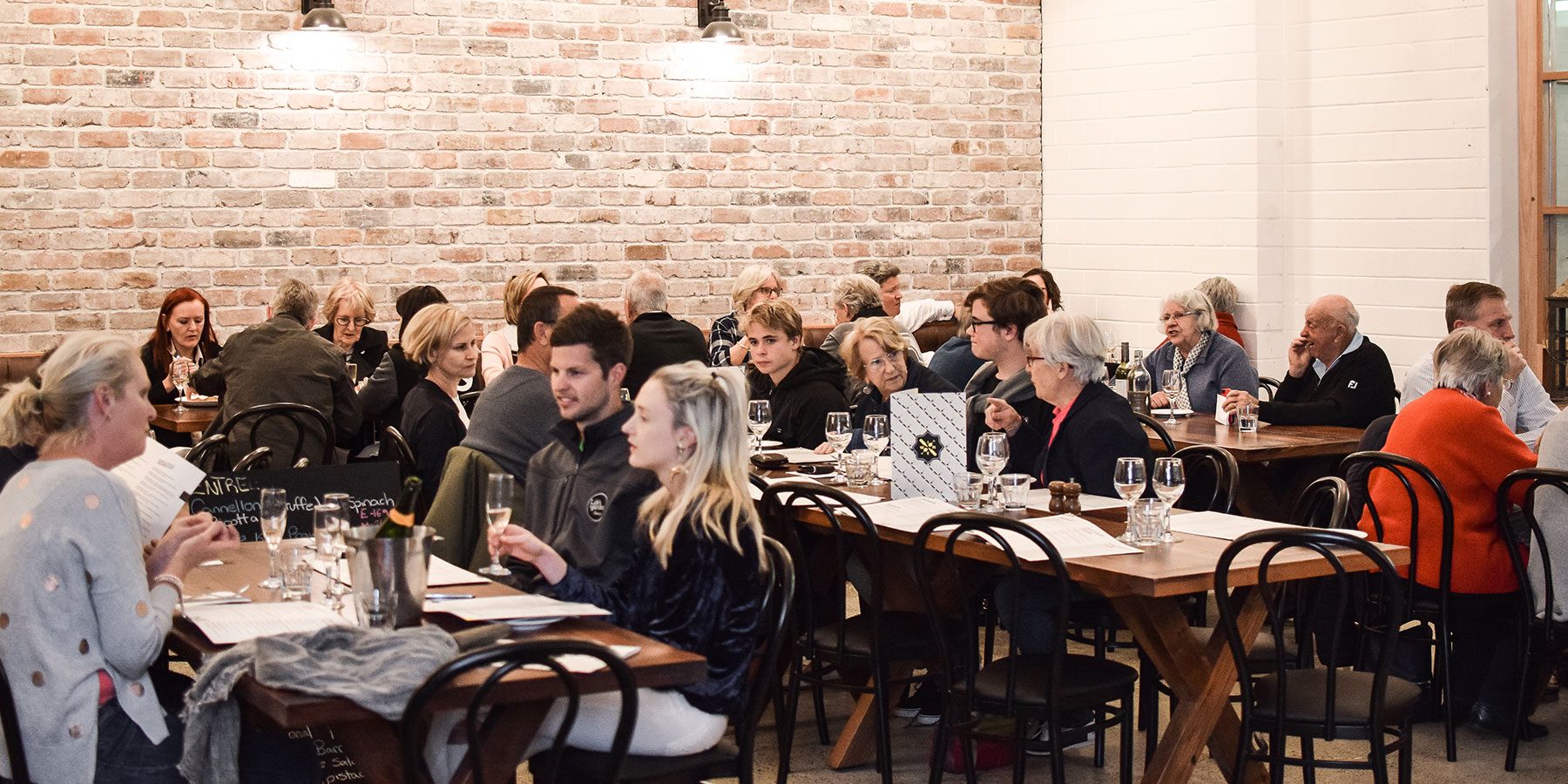 A full dining room at Lucky and Pep's, with families and friends at long timber tables against a white painted brick wall